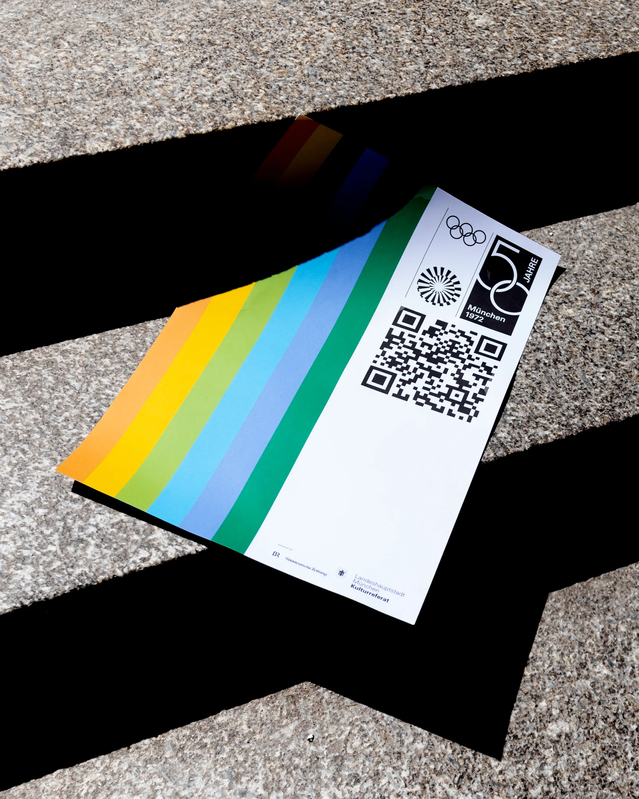 Auf einer Steintreppe liegt ein Flyer des 50-jährigen Jubiläums der Olympischen Spiele in München mit bunten Streifen