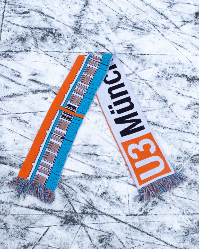 Orange-blauer Fußballlschal, der auf zugrfrorenem Kanal liegt. In U-Bahn Design der Schriftzug "U3 Münchener Freiheit"