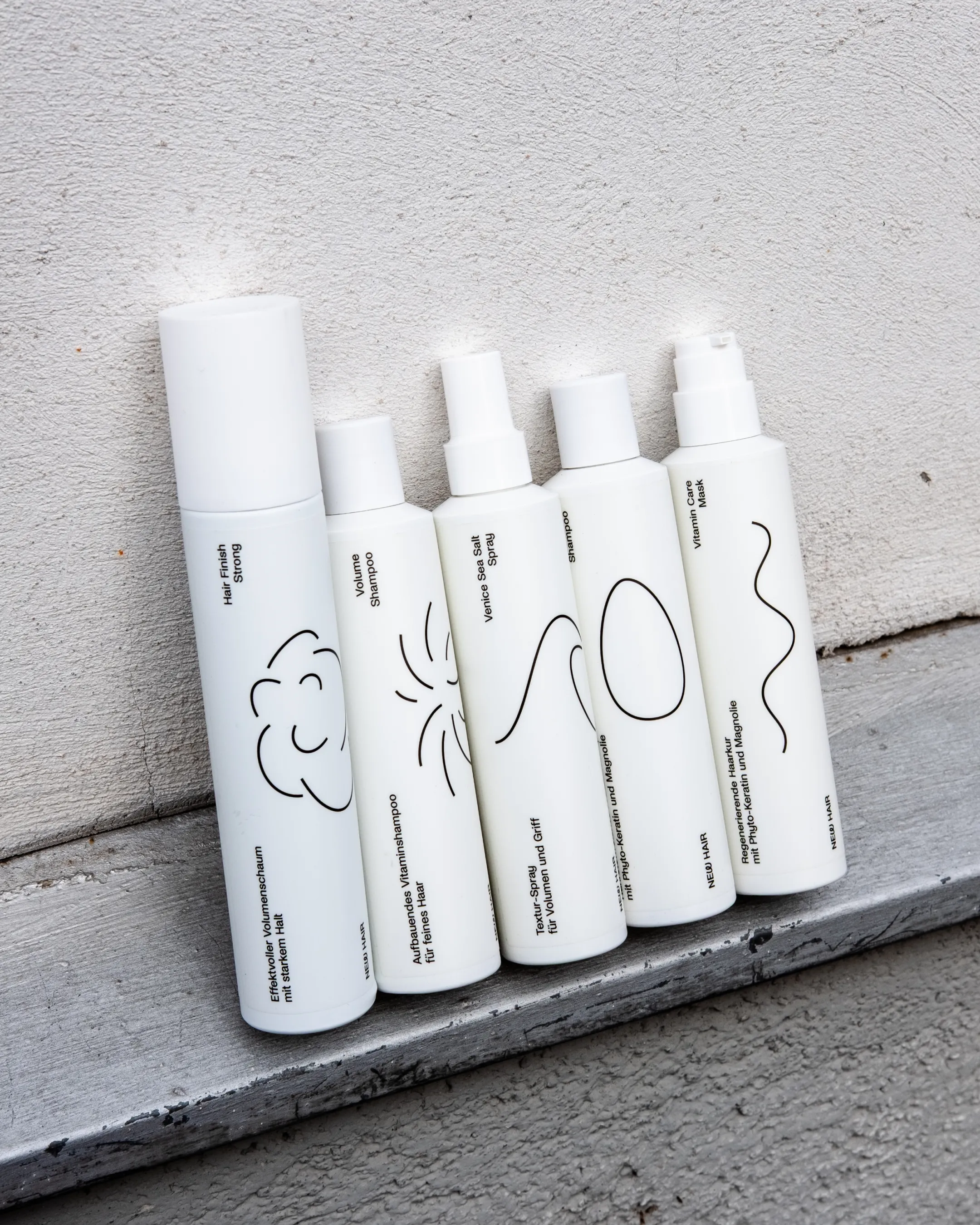 Verschiedene weiße Haarpflegeprodukte mit minimalistischen Line-art Illustrationen und kleiner Typografie lehnen an Hauswand