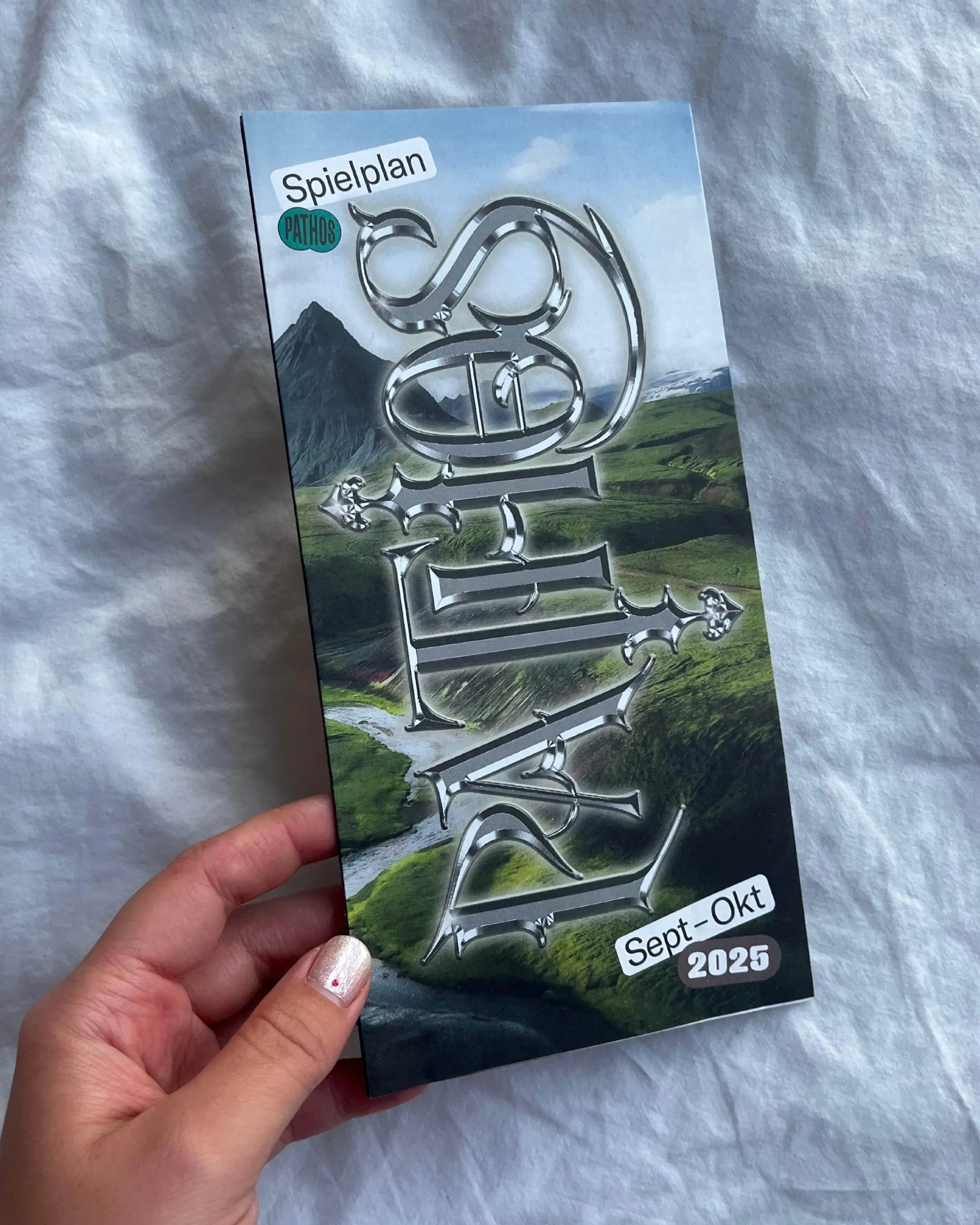 Handyfoto einer Programmflyer-Vorderseite mit den Pathos-Schriftzug in Metall-Mittelalter-Stil auf grüner Berglandschaft