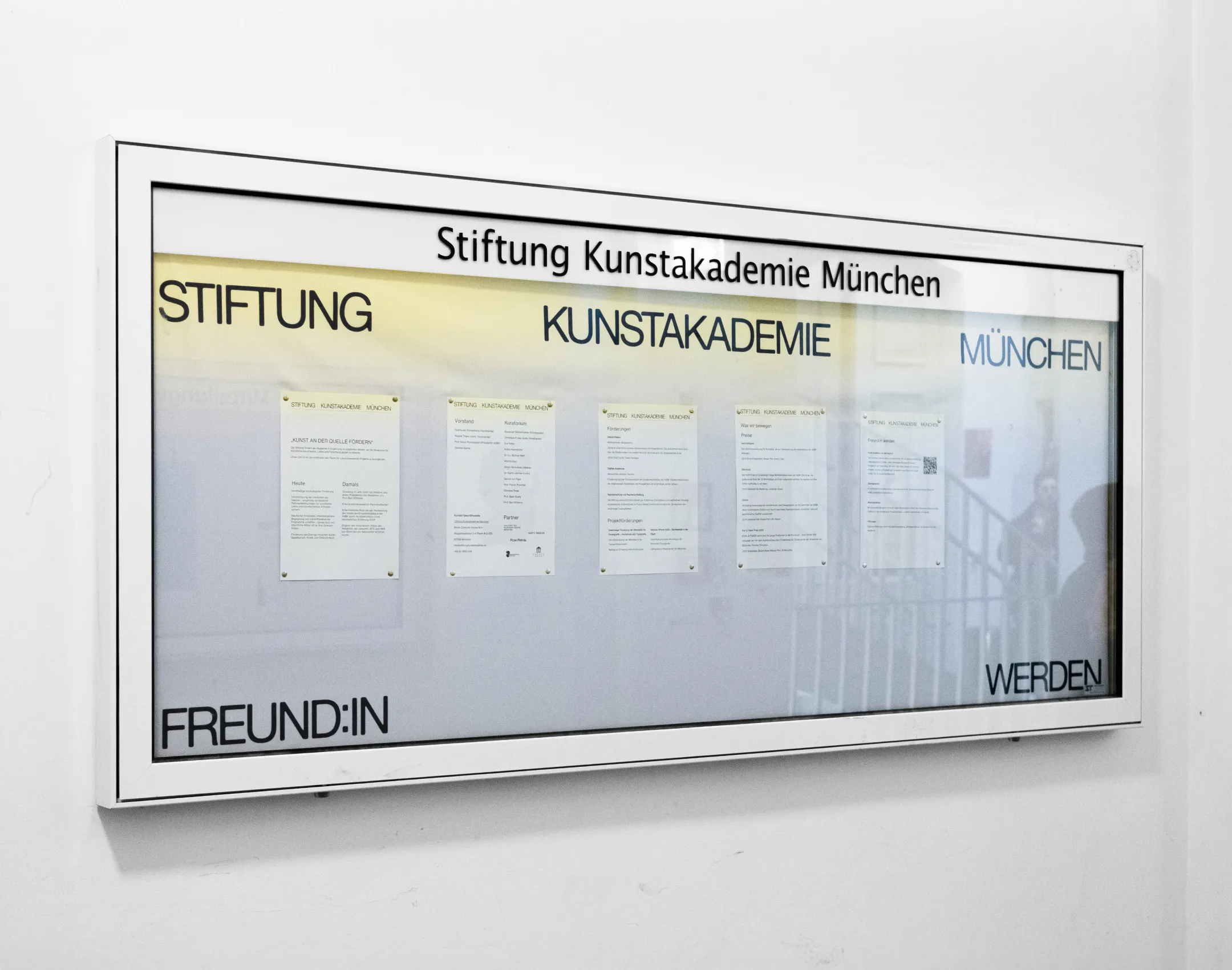 Schaukasten der Stiftung Kunstakademie München mit grau-gelbem Farbverlauf als Hintergrund und Logo im Flur der Akademie